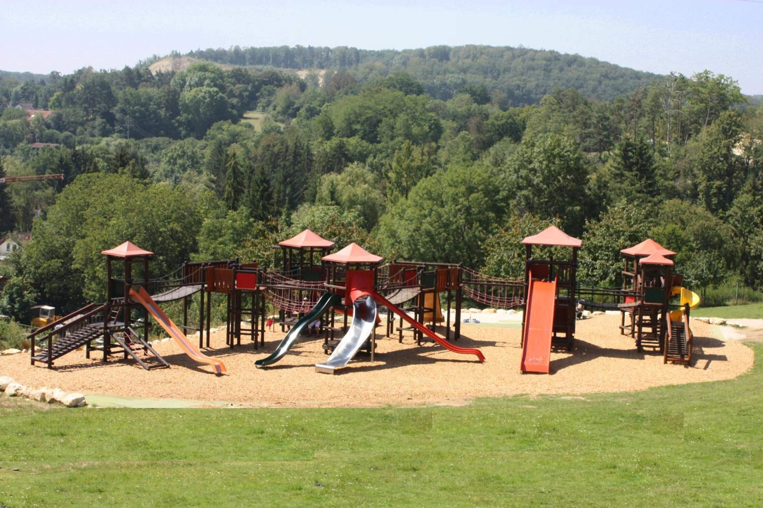 A playground with multiple slides and climbing structures, surrounded by a grassy area and a forested hill in the background.