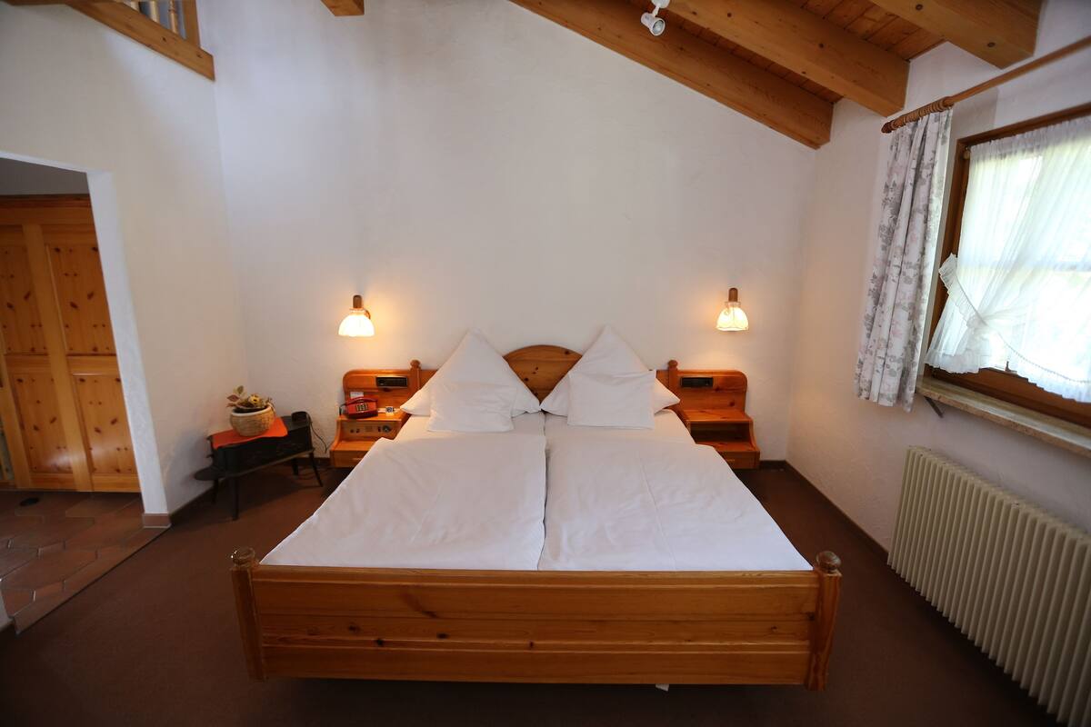 A wooden bed in a room with white walls, two bedside tables with lamps, a window with curtains, and a radiator.