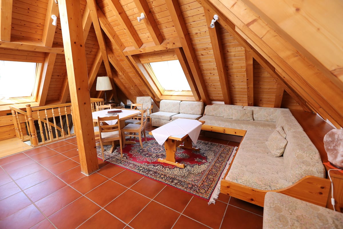A cozy attic bedroom with a wooden ceiling, a sofa, a small table, and a chair.