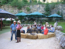 Un grupo de personas sentadas alrededor de una mesa circular con fardos de heno, bajo sombrillas verdes, en un espacio al aire libre con un muro de piedra y vegetación de fondo.