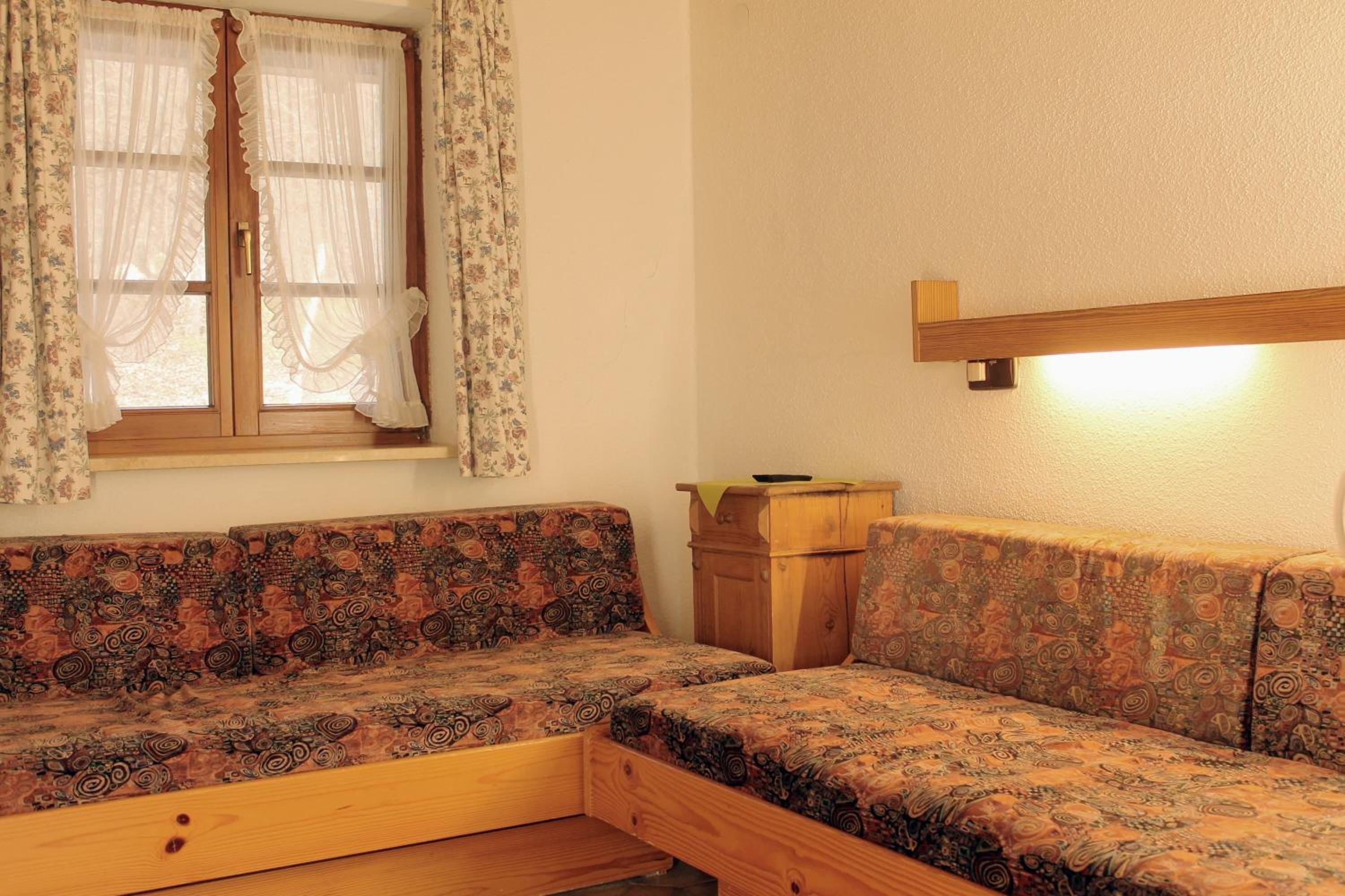Una stanza accogliente con un divano letto in legno, un piccolo mobiletto in legno e una finestra con tende floreali.