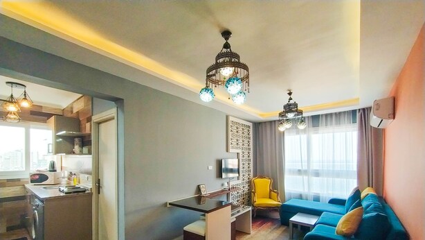 Een moderne woonkamer met een blauwe bank, een klein tafeltje en een gele stoel. Er is een keukenhoek met een magnetron en een spoelbak. De kamer heeft houten vloeren en een kroonluchter.