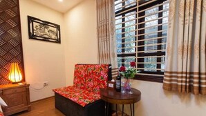 A room with a wooden floor, a floral-patterned chair, a small table with a vase of flowers, a lamp, and a window with curtains. - Dollar Homestay & Travel (Hanoi)