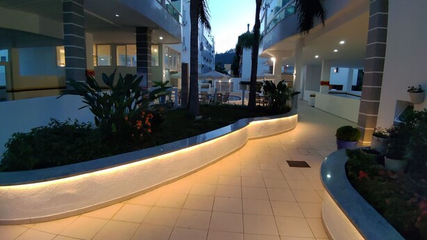 A modern hotel corridor with a curved walkway, outdoor seating area, and potted plants. - Porto Del Mar - Canasvieiras (Florianópolis)