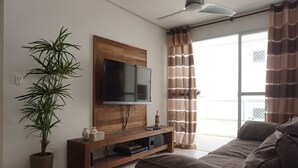A modern living room with a flat-screen TV mounted on a wooden panel, a large potted plant, a sofa, and a sliding glass door with curtains. - Porto Del Mar - Canasvieiras (Florianópolis)