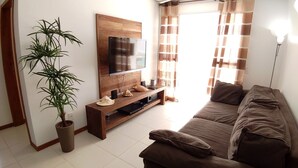 A living room with a wooden TV stand, a flat-screen TV, a brown sofa, a potted plant, and a window with curtains. - Porto Del Mar - Canasvieiras (Florianópolis)