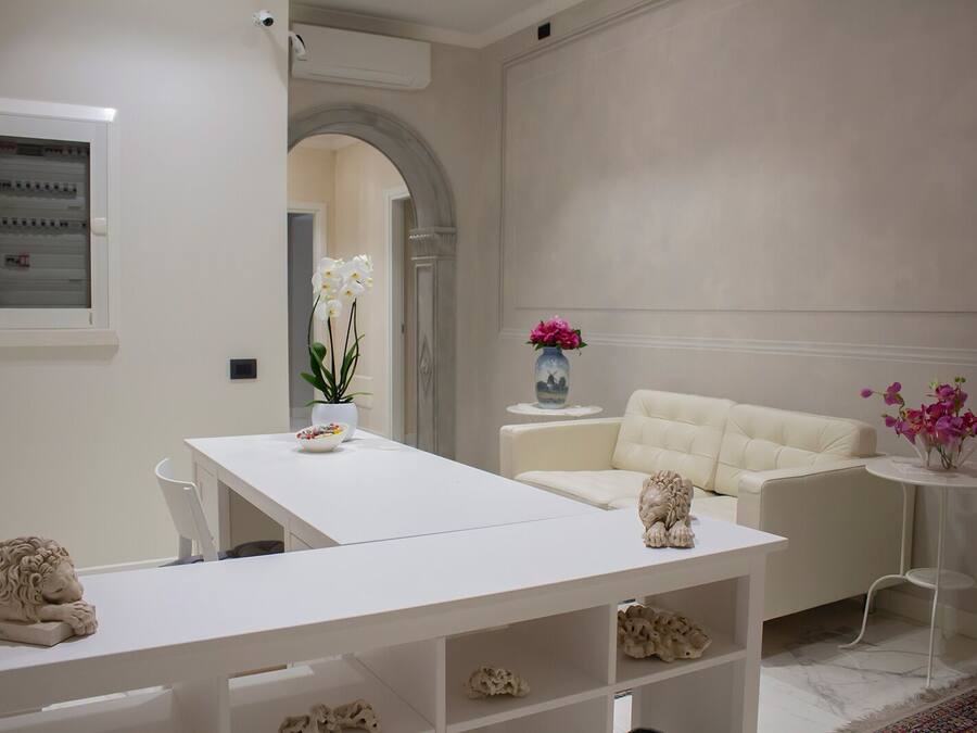 A modern, minimalist room with a white dining table, a white sofa, and a white armchair. There are decorative items like a vase with flowers and a sculpture on the table.