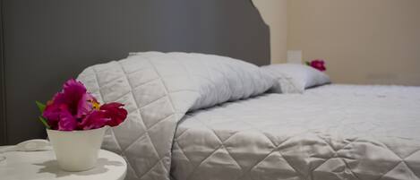 Una cama con una colcha acolchada y una almohada blanca. Hay una mesita de noche blanca con una maceta blanca que contiene flores rosas.