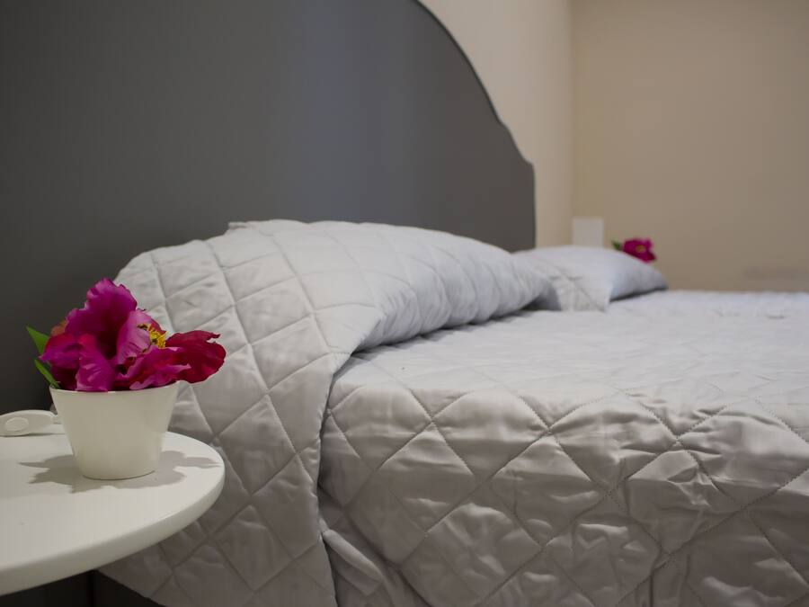 Un lit avec une couvre-lit matelassé et un oreiller blanc. Il y a une table de chevet blanche sur laquelle se trouve un pot blanc contenant des fleurs roses.