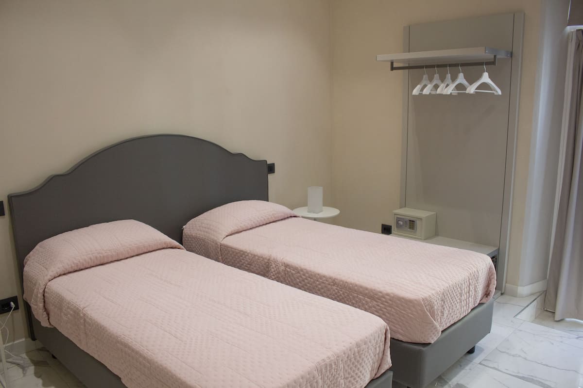 Quarto com duas camas, uma mesinha de cabeceira, um espelho e um guarda-roupa com roupas penduradas.
