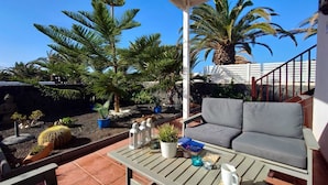 Un patio al aire libre con un sofá gris, una mesa con libros y una vela, y diversas macetas con plantas.