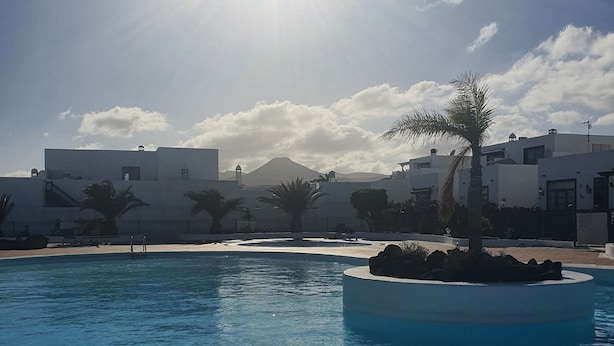 Una piscina con superficie de agua azul cristalina, rodeada por un solárium blanco. Al fondo, edificios blancos y una montaña a lo lejos.