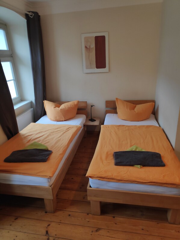 A room with two single beds, each with an orange blanket and a pillow. There is a window with curtains, a nightstand with a lamp, and a framed picture on the wall. - Altstadt Pension Potsdam (Potsdam)