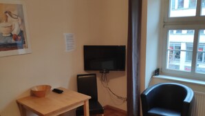 A room with a wooden floor, a table with chairs, a black chair, a television mounted on the wall, and a window with a view of buildings outside. - Altstadt Pension Potsdam (Potsdam)