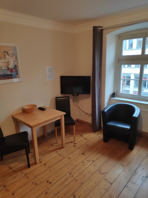Una habitación con piso de madera, una mesa con sillas, una silla negra, un televisor montado en la pared y un ventanal con vistas a edificios.