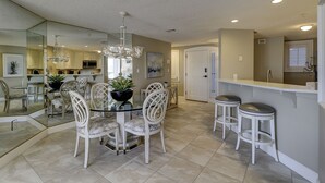 A modern kitchen with a glass dining table, white chairs, a chandelier, and a white door. - 3209 Windsor Court South - Beautiful Panoramic Oceanfront Views! (Hilton Head Island)