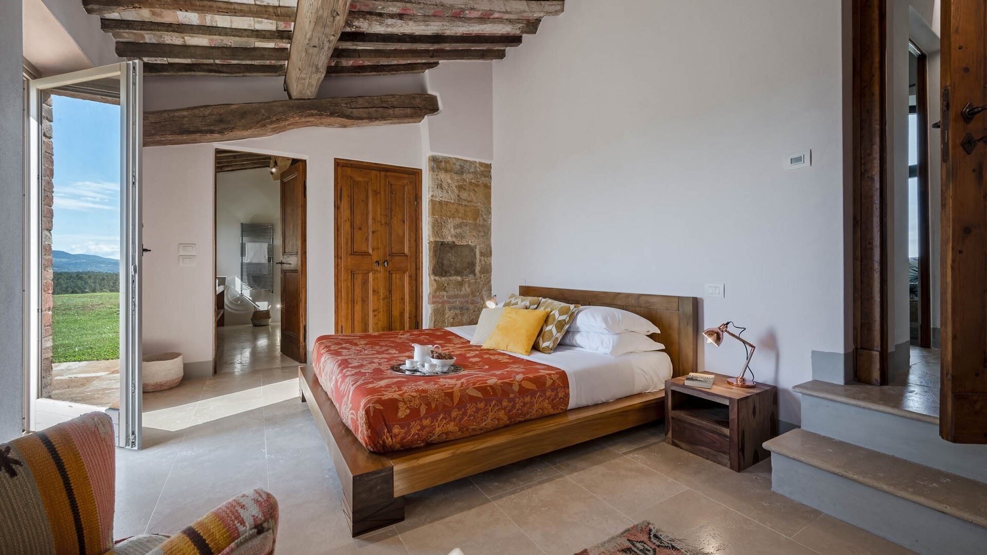 A bedroom with a large bed, wooden beams, a stone wall, and a view of the outdoors.