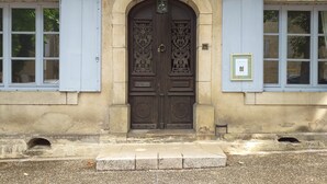 Ein Gebäude mit einer großen, verzierten Holztür, zwei Fenstern mit Rollläden und einer kleinen Wandplakette.
