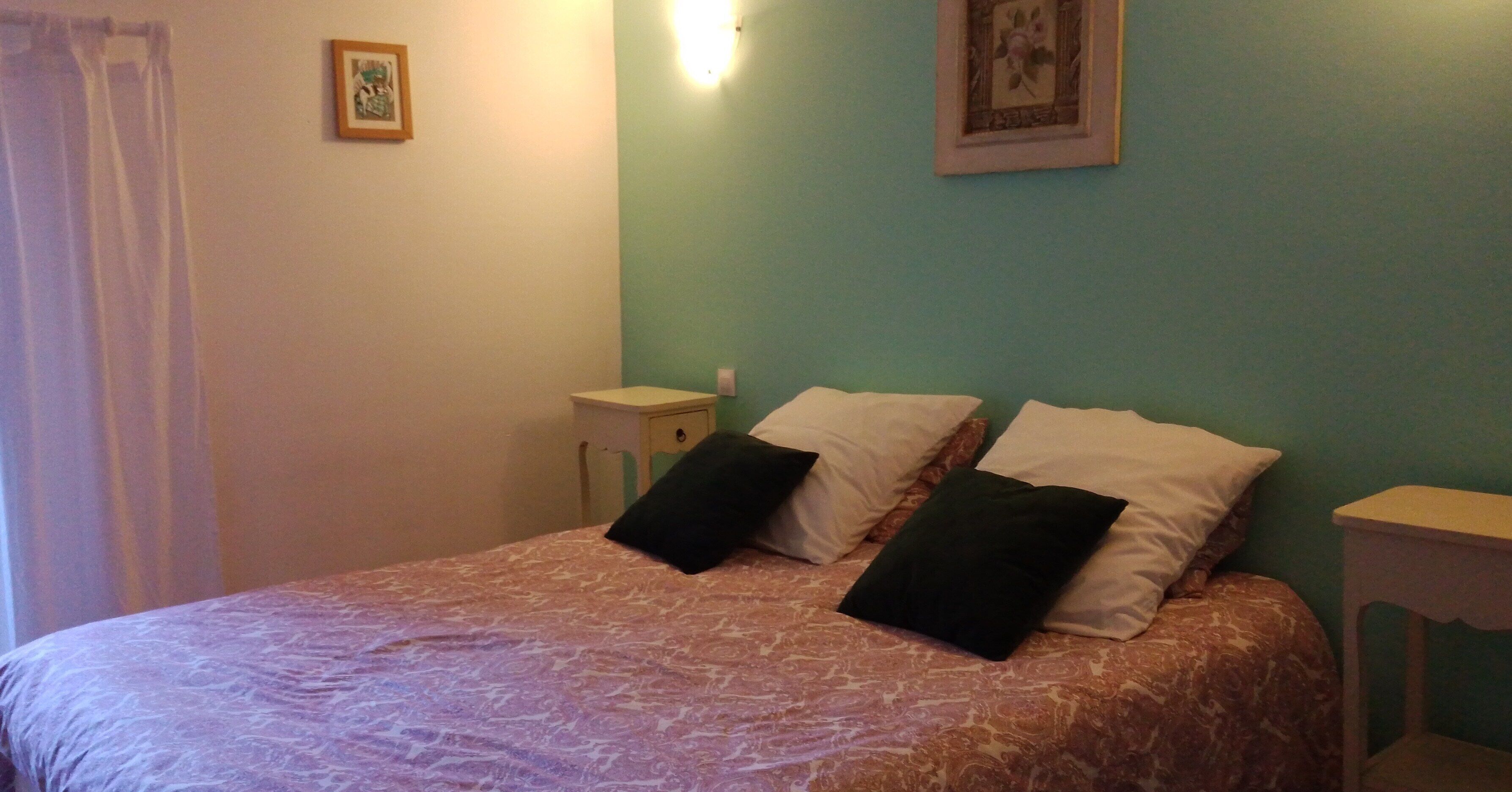 A bedroom with a bed, two pillows, a nightstand, a wall-mounted light, and a framed picture.