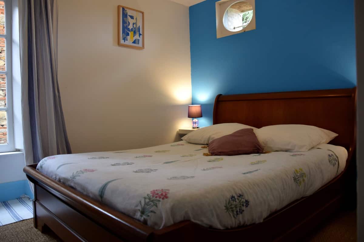 Een slaapkamer met een houten bed, een nachtkastje met een lamp, een raam met gordijnen en een wandklok.