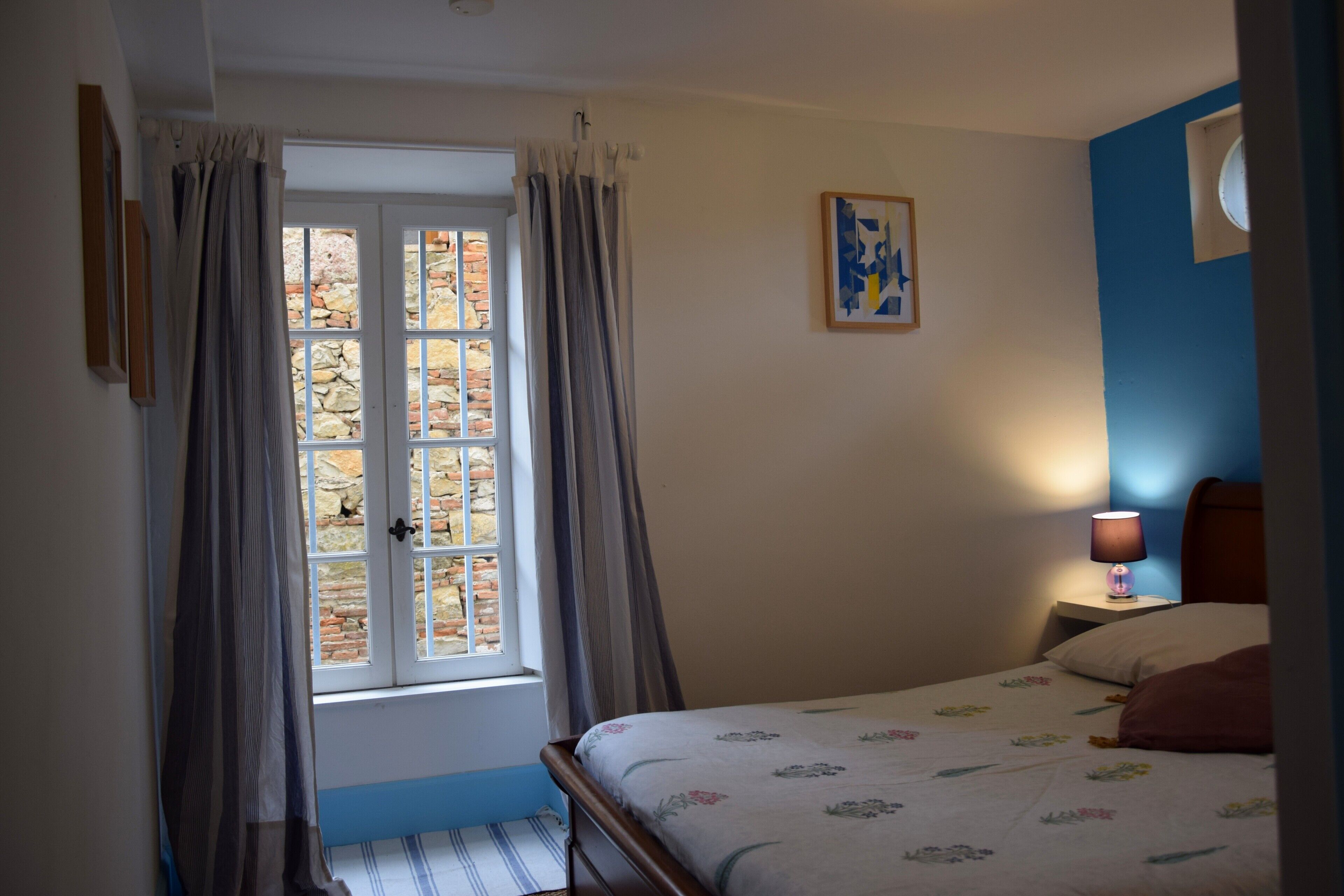 Une chambre à coucher avec un lit, une table de chevet, une lampe, une fenêtre avec des rideaux et un tableau au mur.