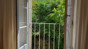 Eine offene Schiebeglastür führt zu einem Balkon mit Metallgeländer, einer Steinmauer und Blick auf Grünflächen und Hügel.