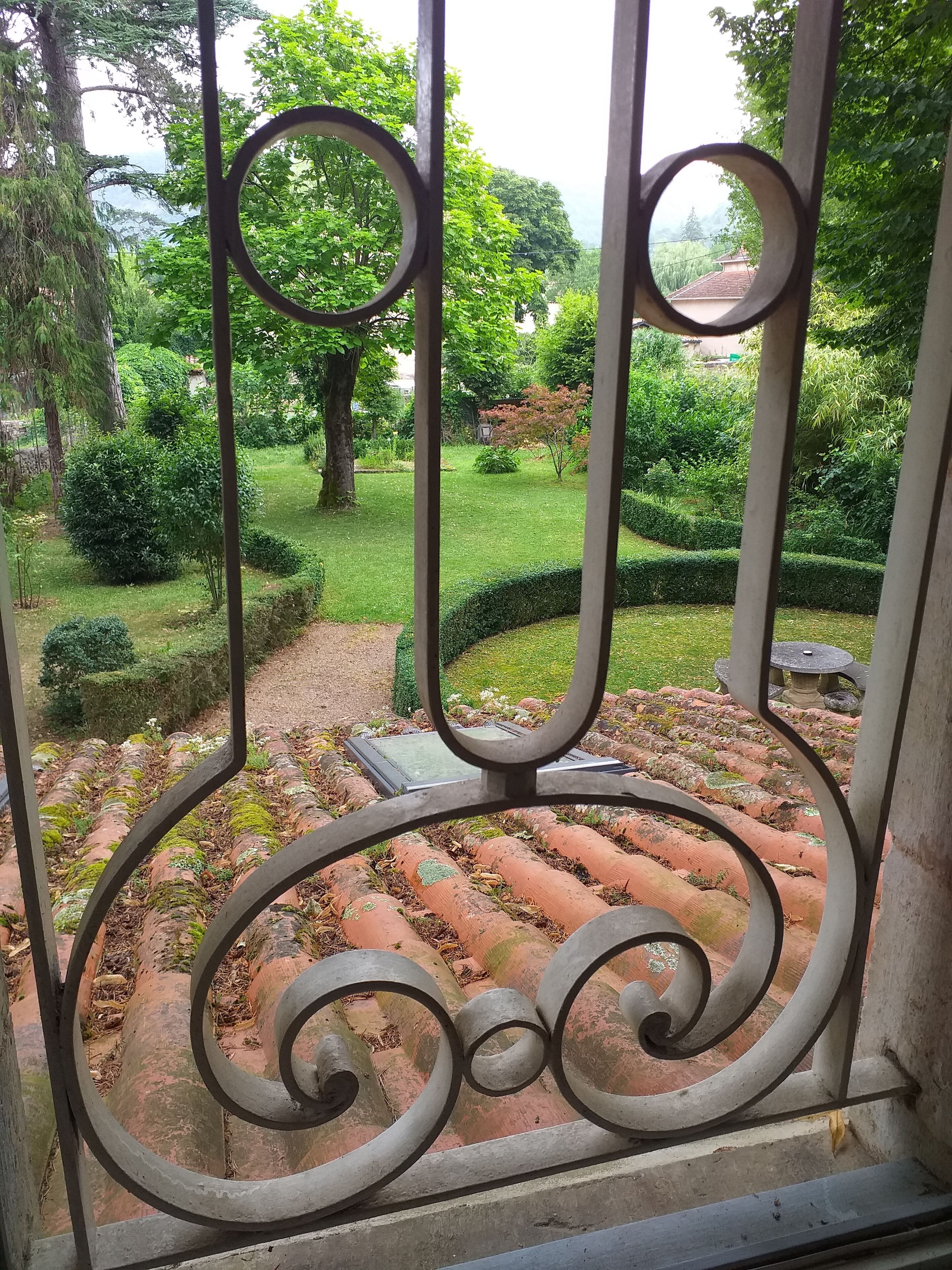 Vista de un jardín a través de una reja metálica, con un camino de piedra y abundante vegetación.