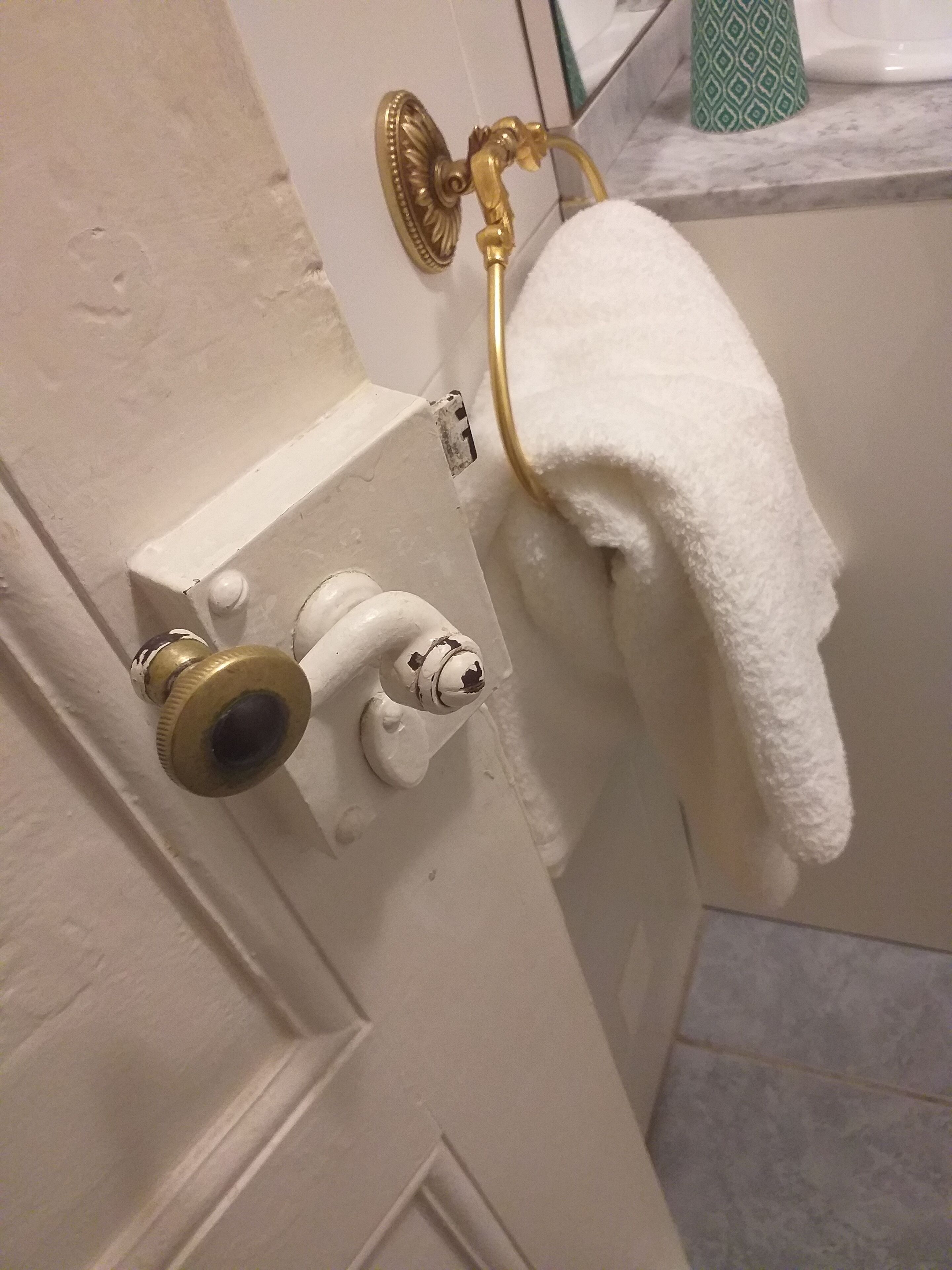 Un baño con una puerta blanca, una toalla colgada en un toallero dorado y un recipiente verde con estampado sobre la encimera.