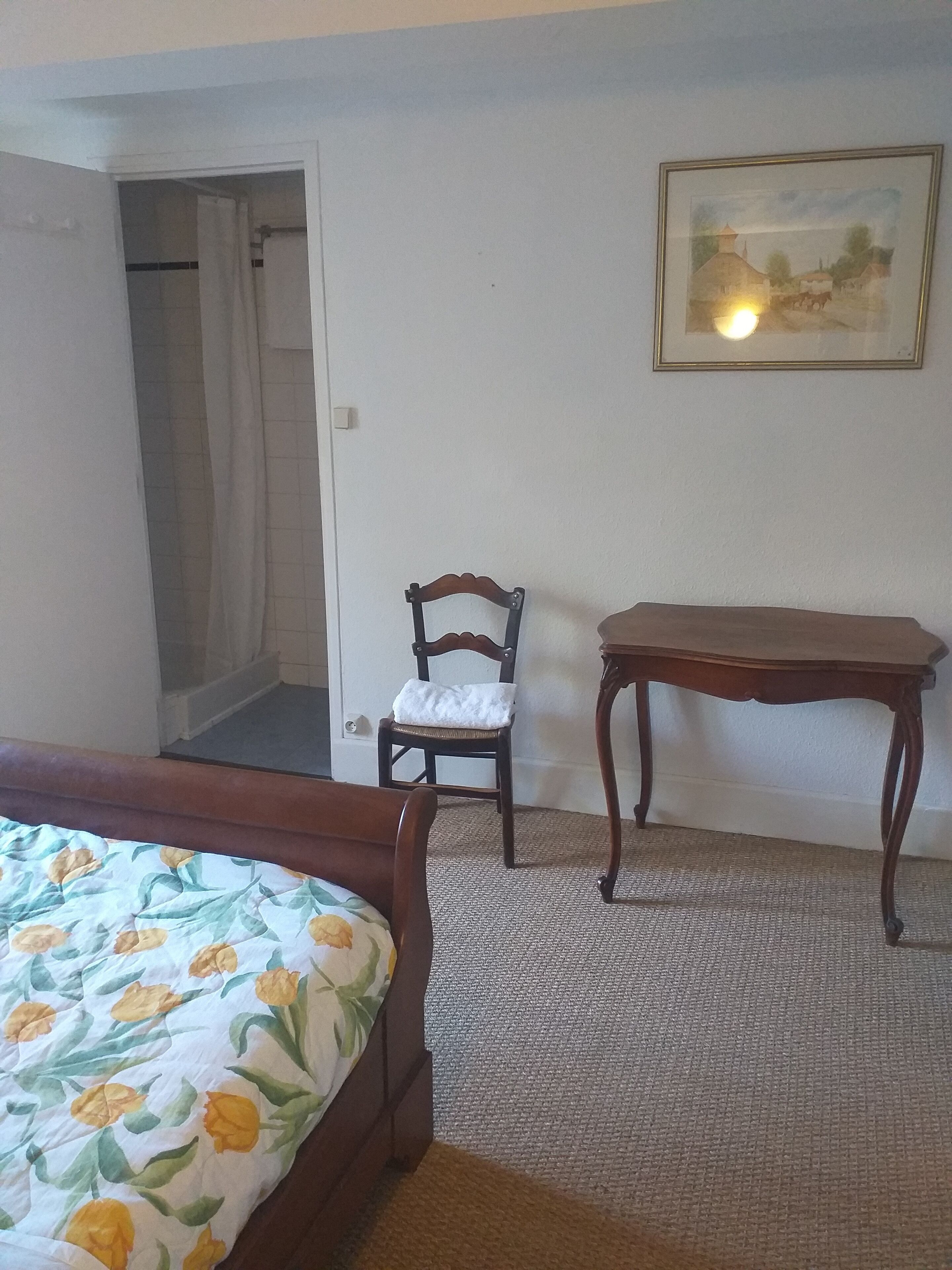 A bedroom with a bed, a wooden chair, a small table, and a painting on the wall.