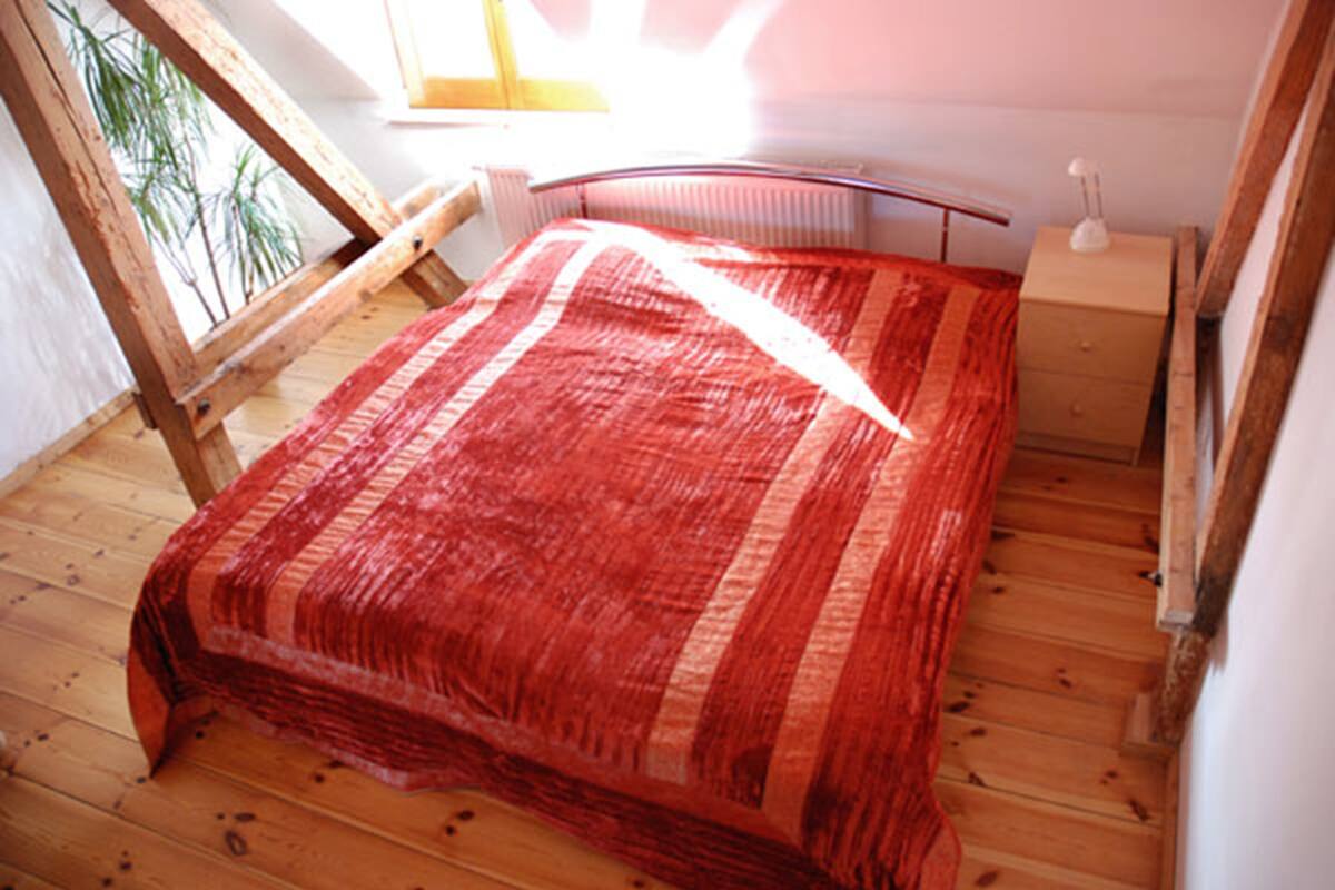 Una habitación con piso de madera, una cama con manta roja, una mesita de noche de madera y una ventana con vista a árboles.