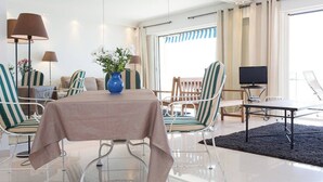 Habitación de hotel con mesa de vidrio, sillas a rayas, televisor y ventana grande con cortinas.