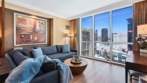A modern living room with a large window offering a city view, a grey sofa with a blanket, and a coffee table with glasses. - Luxury StripView 2-King Suites+No Resort Fee+Pool (Las Vegas)