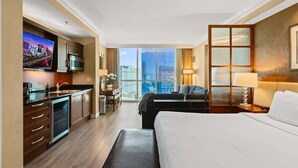 A hotel room with a bed, a desk, a television, a kitchenette, and a large window offering a city view. - Luxury StripView 2-King Suites+No Resort Fee+Pool (Las Vegas)