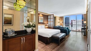 A modern hotel room with a large bed, a seating area with a sofa and armchairs, a dining table, and a view of the cityscape. - Luxury StripView 2-King Suites+No Resort Fee+Pool (Las Vegas)