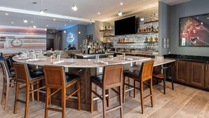 A modern bar area with a central island, bar stools, and a variety of bottles and glasses. - Cambria Hotel Nashville Airport (Nashville)