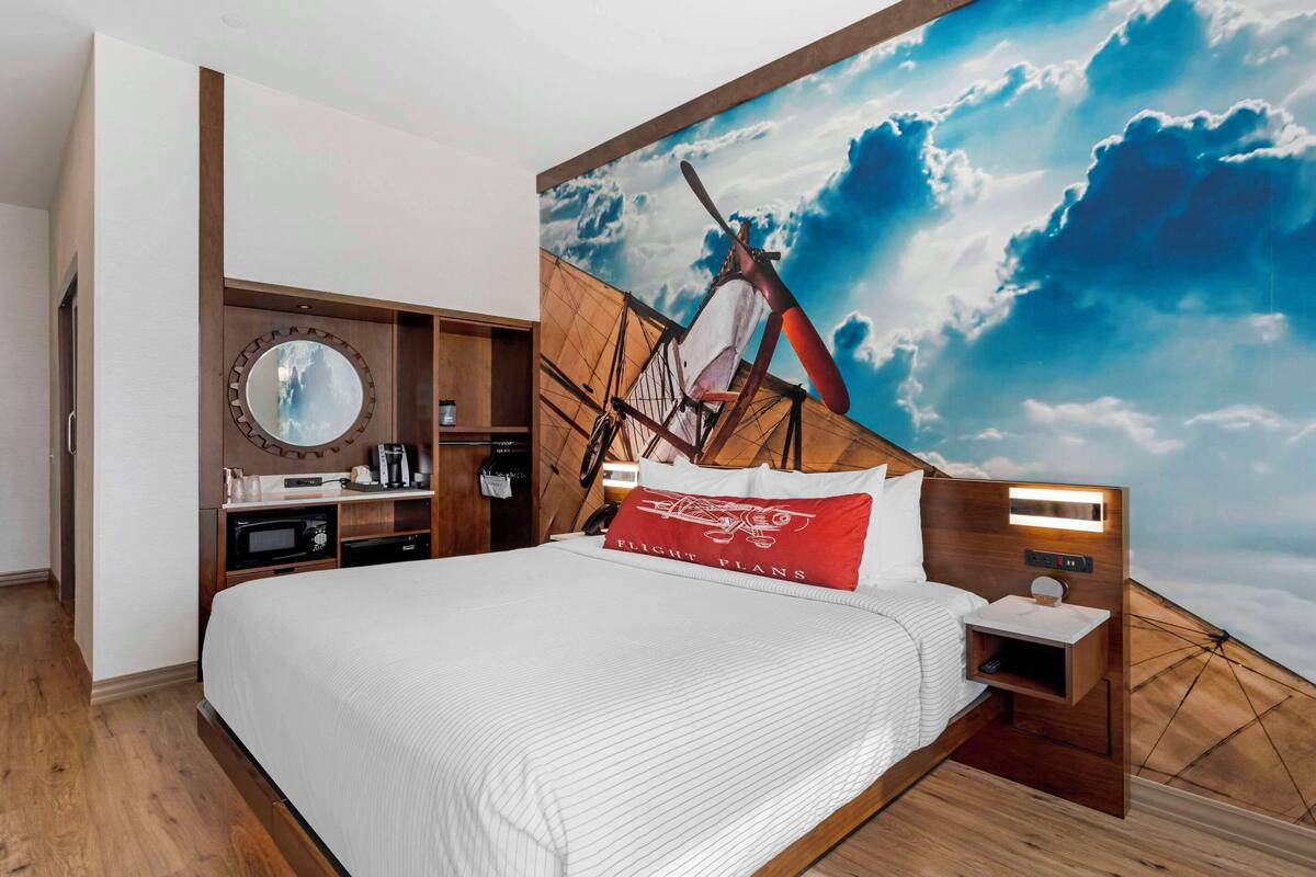 Une chambre d’hôtel avec un grand lit, une tête de lit en bois, une fresque murale représentant un navire, un miroir rond, une table de chevet avec une lampe, et une étagère intégrée contenant une machine à café et d’autres objets.