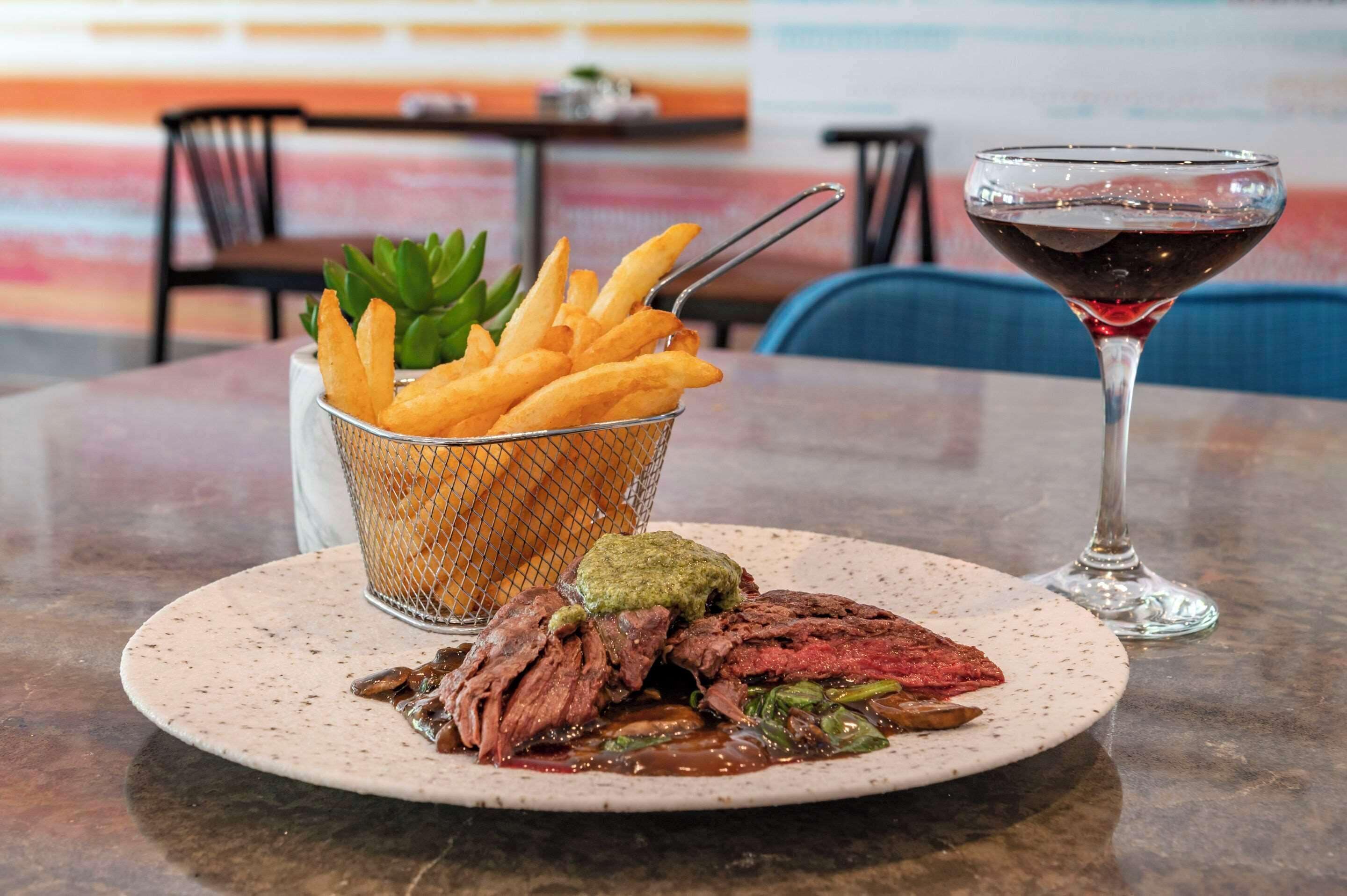 Ein Teller mit Steak, Pommes und grüner Soße, dazu ein Glas Rotwein auf einem Tisch.