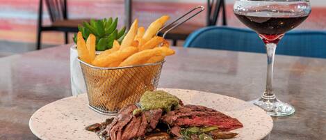 Une assiette contenant un steak, des frites et une sauce verte, accompagnée d’un verre de vin rouge, le tout posé sur une table.