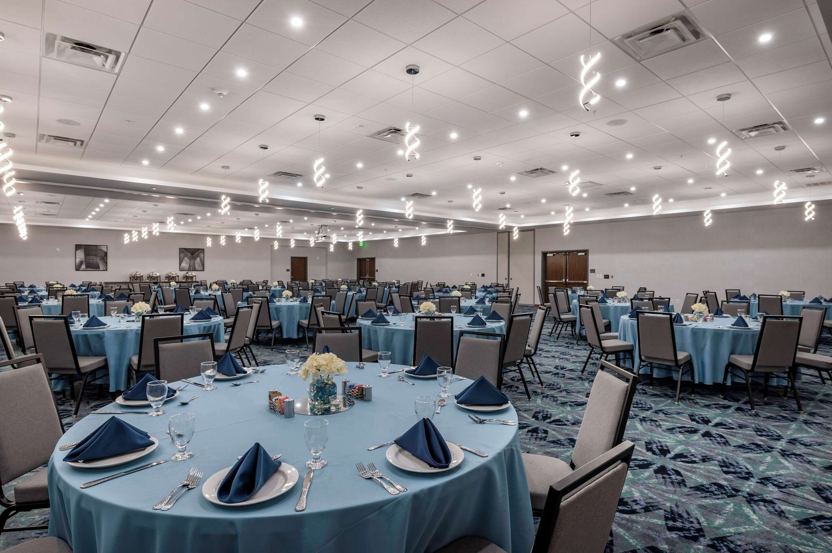 Un amplio salón de banquetes con mesas redondas dispuestas para el servicio de comidas, equipado con sillas, platos, cubiertos y copas.