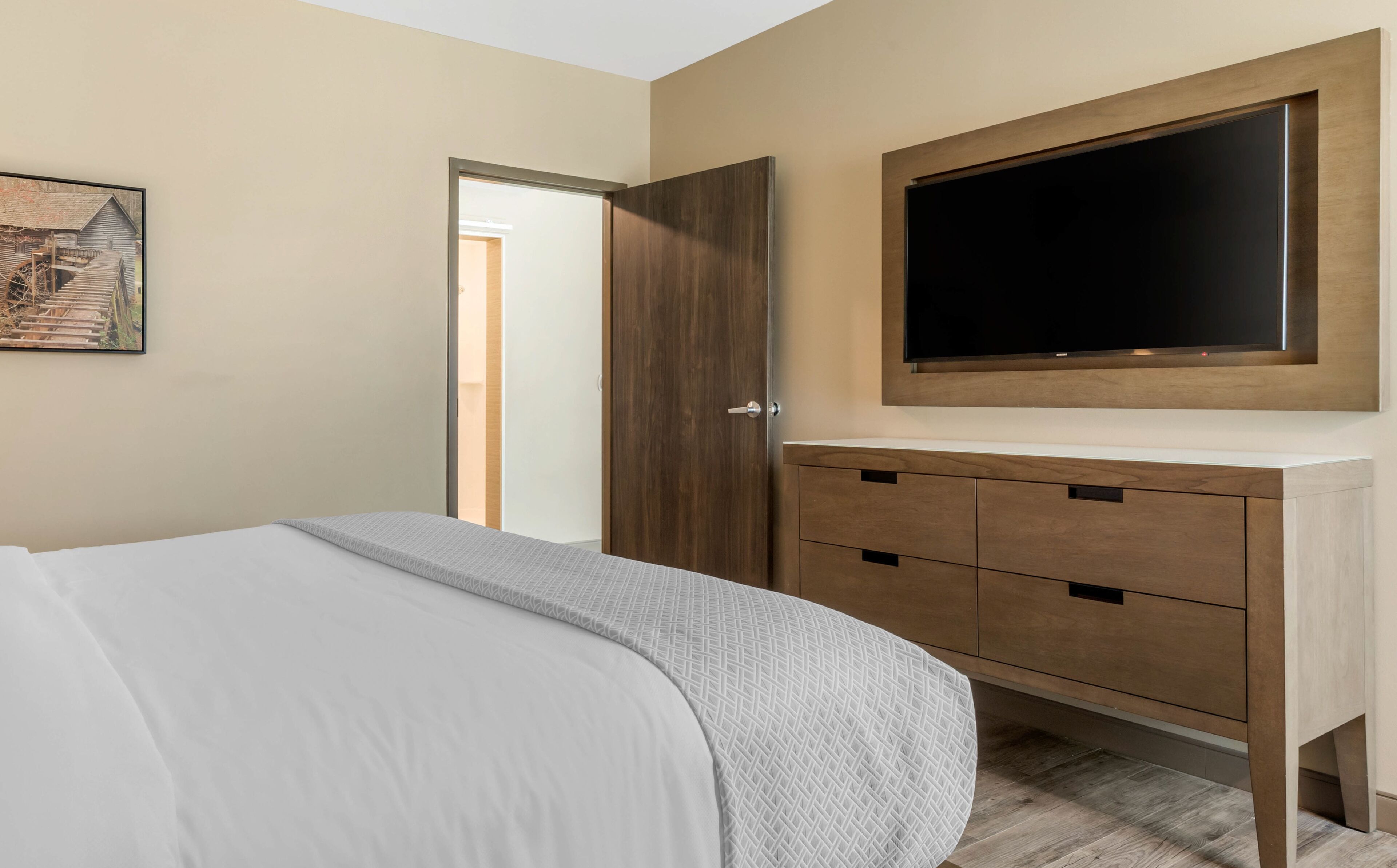 A bedroom with a bed, a wooden wardrobe, a television mounted above it, and a painting on the wall.