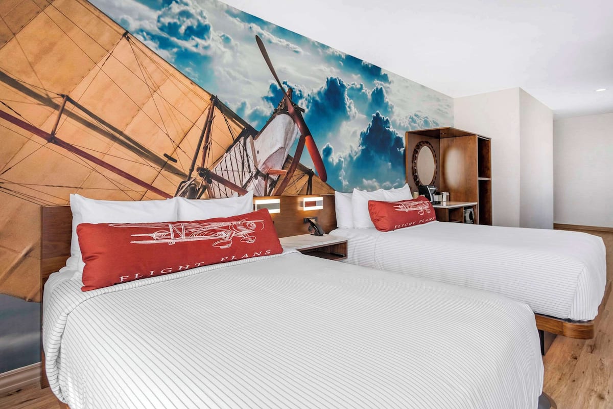 Une chambre d’hôtel avec deux lits, chacun agrémenté de draps blancs et d’oreillers rouges, une grande fresque murale représentant un voilier et une table de chevet en bois avec un miroir.