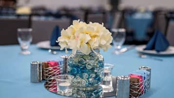 Een gedekte tafel met een glazen pot gevuld met blauwe en witte decoratieve items, omringd door zilveren kandelaars en een klein mandje met papieren.