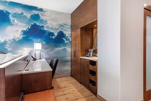 Habitación compacta con escritorio de madera, una silla, un mural grande de nubes en la pared y un mueble empotrado con espejo y máquina de café.