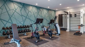 Ein Fitnessraum mit Trainingsgeräten, einer Bank, Kurzhanteln und einer Wand mit geometrischen Mustern.