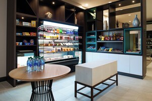 Un espacio comercial moderno con estanterías llenas de diversos productos, un expositor refrigerado y una zona de estar con una mesa y un banco.