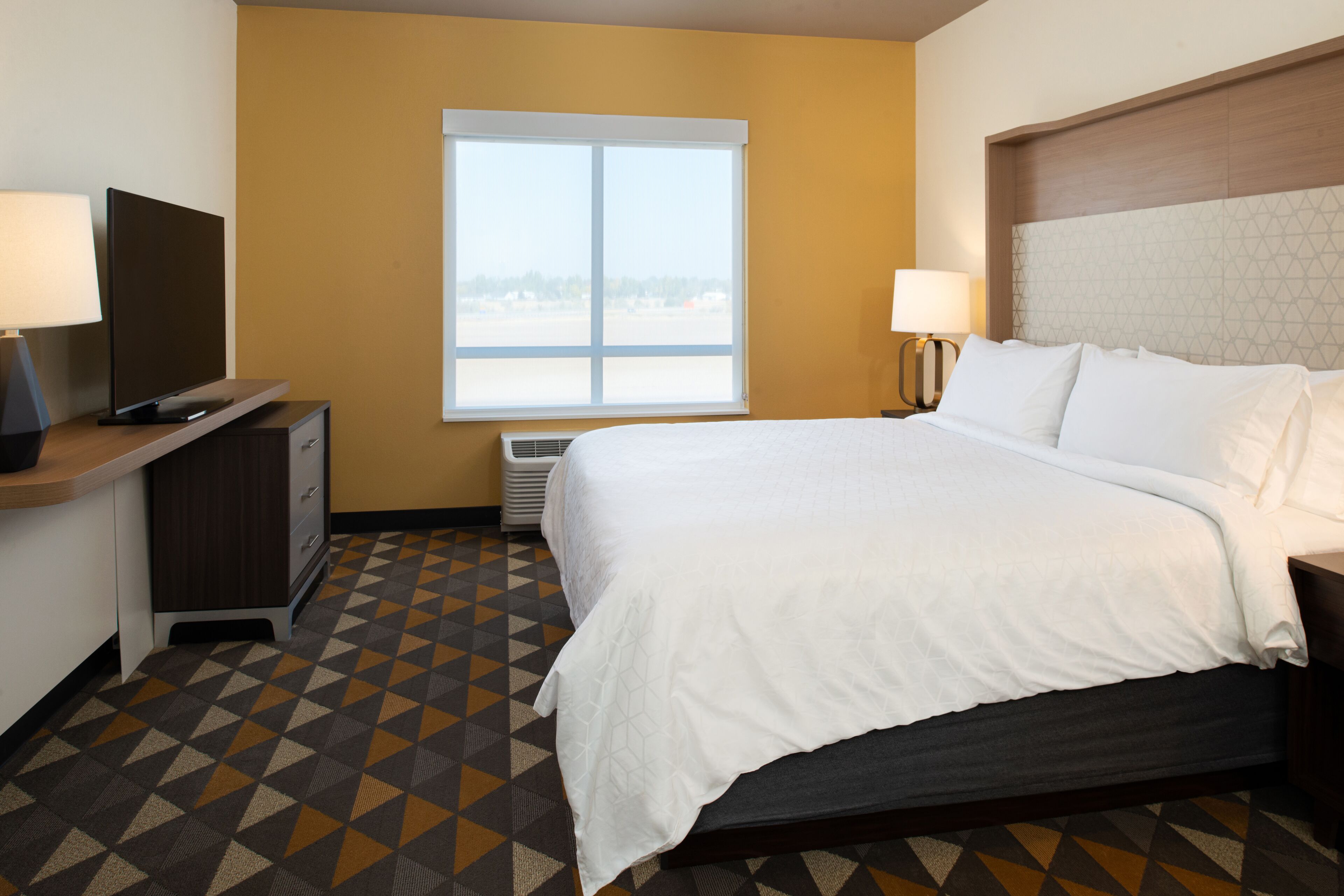 A hotel room with a bed, a desk with a TV, a window, and a patterned floor.
