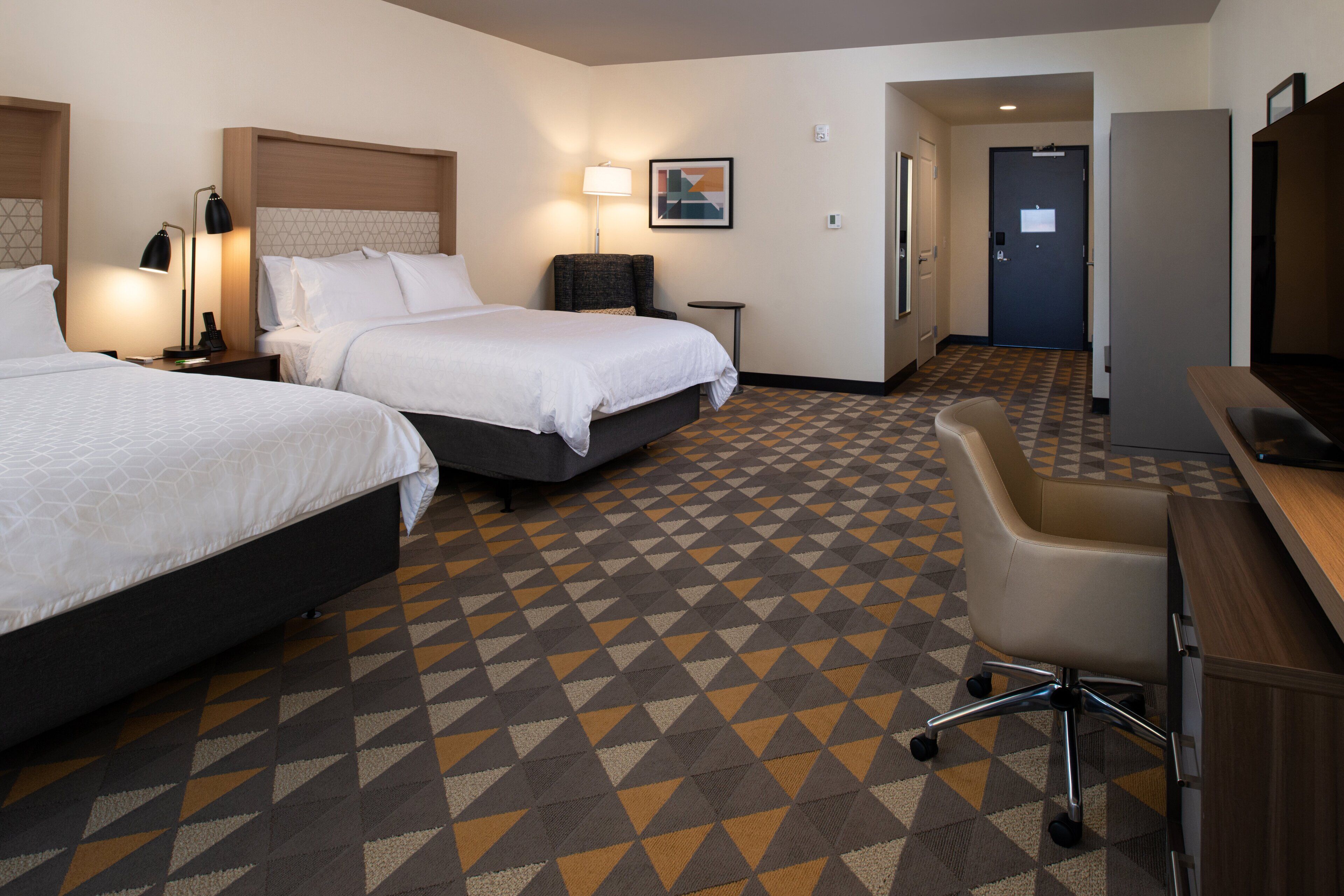 A hotel room with two beds, a desk, a chair, and a patterned carpet.
