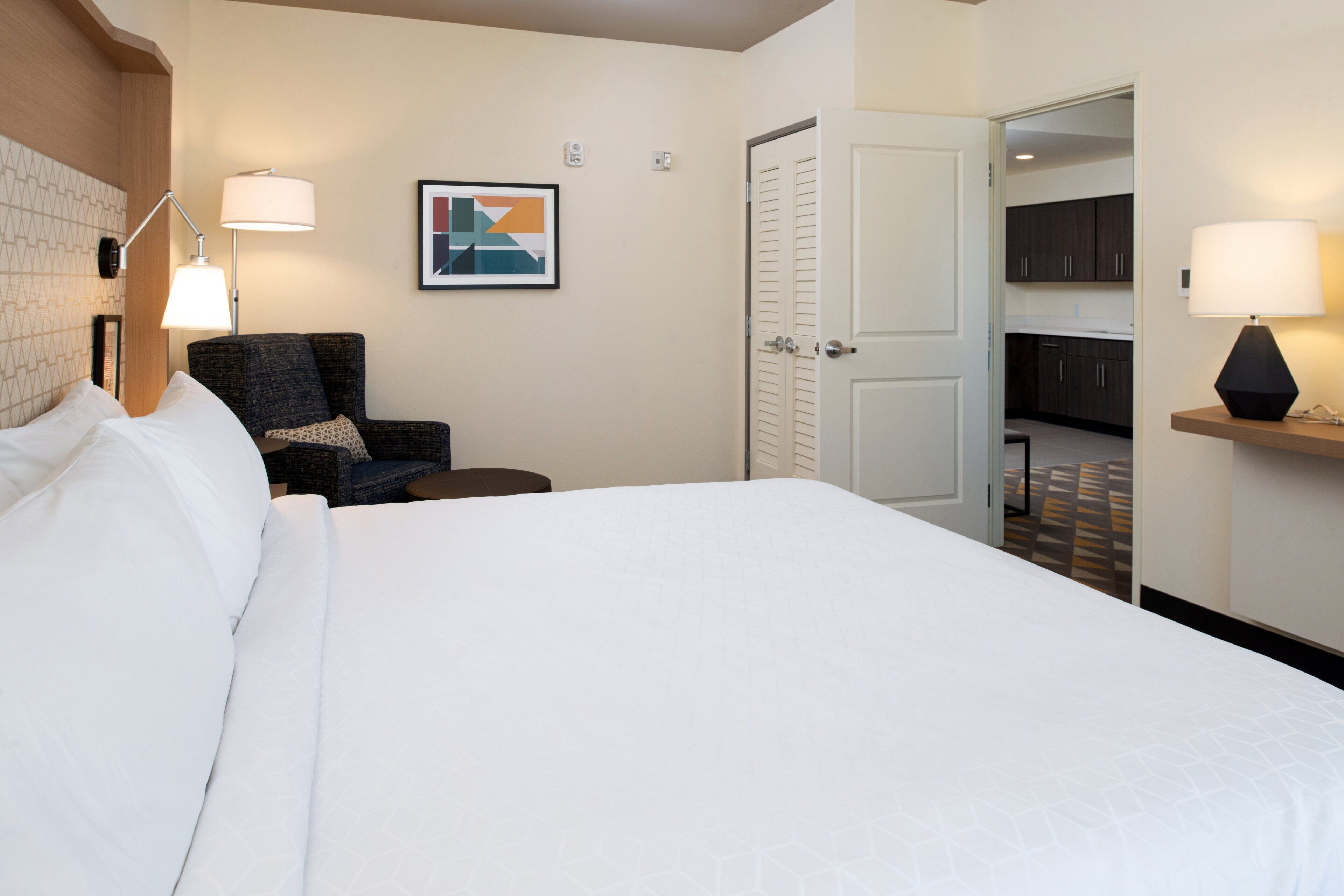A hotel room with a large bed, a chair, a desk, and a door leading to a kitchen area.
