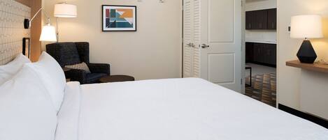 A hotel room with a large bed, a chair, a desk, and a door leading to a kitchen area.