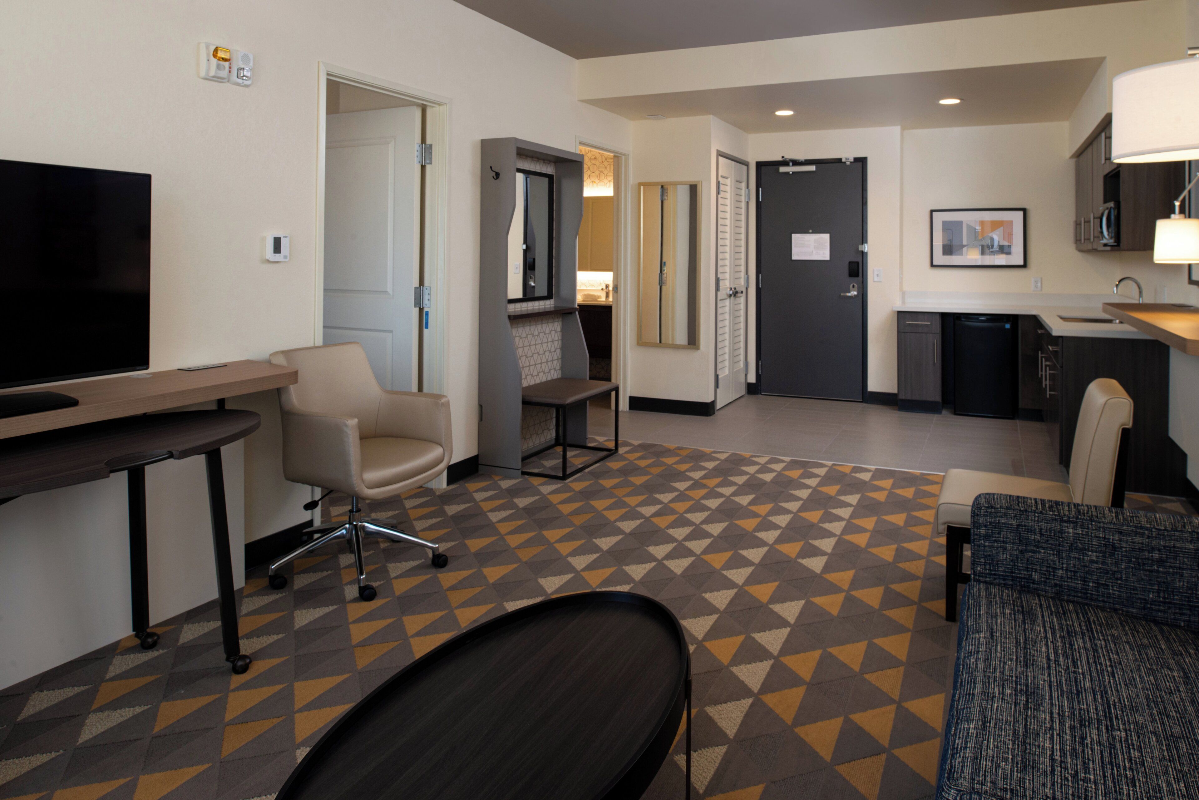 A hotel room with a flat-screen TV, a desk, a chair, a sofa, a kitchen area with a sink and cabinets, and a patterned carpet.
